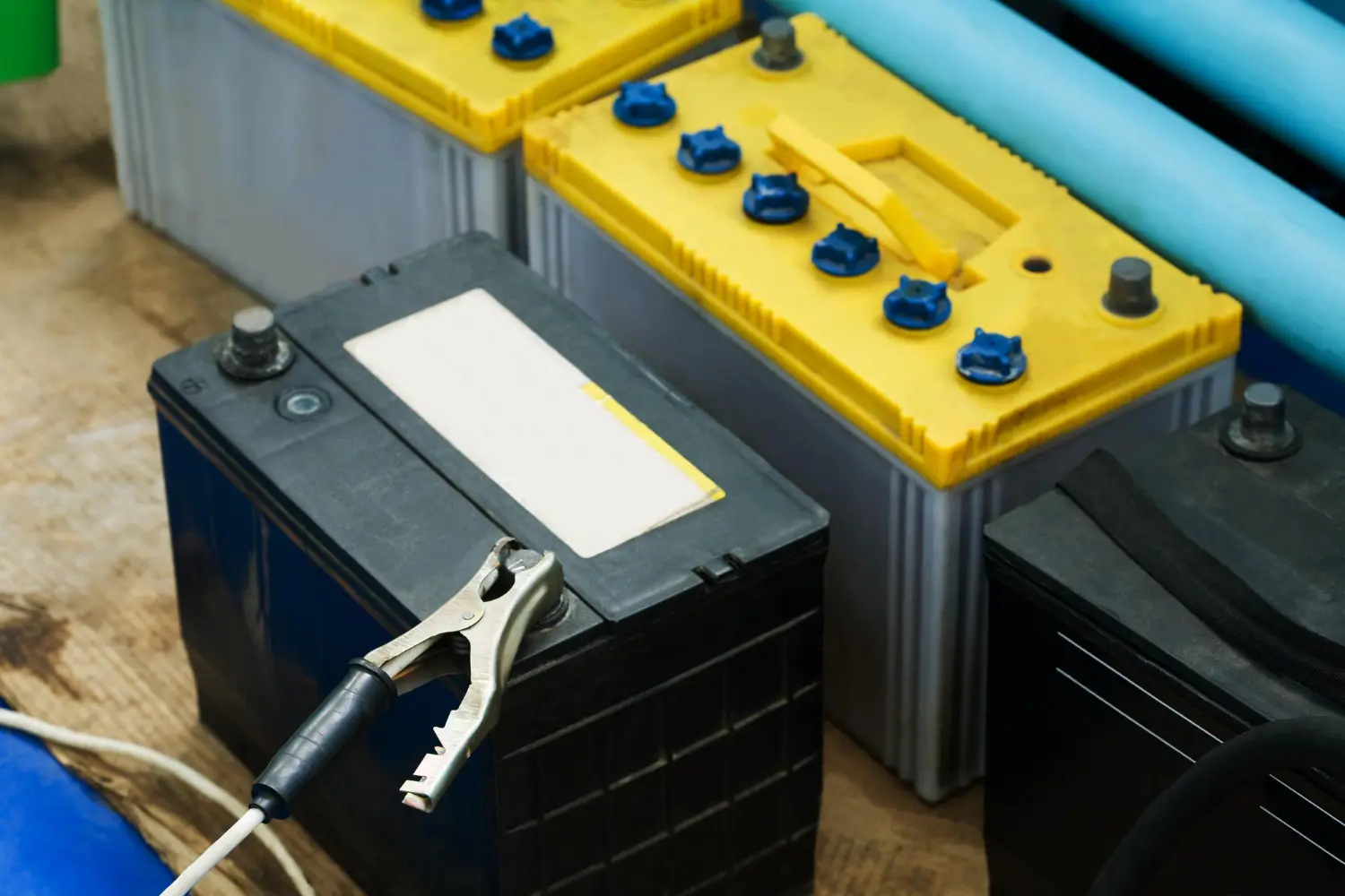 Several car batteries on a wooden surface with a jumper cable connected to one battery.